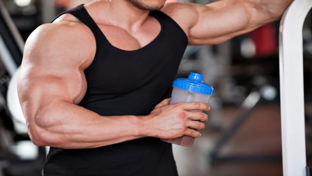 A bodybuilder holding a protein shake casually displaying his 16 inch forearms