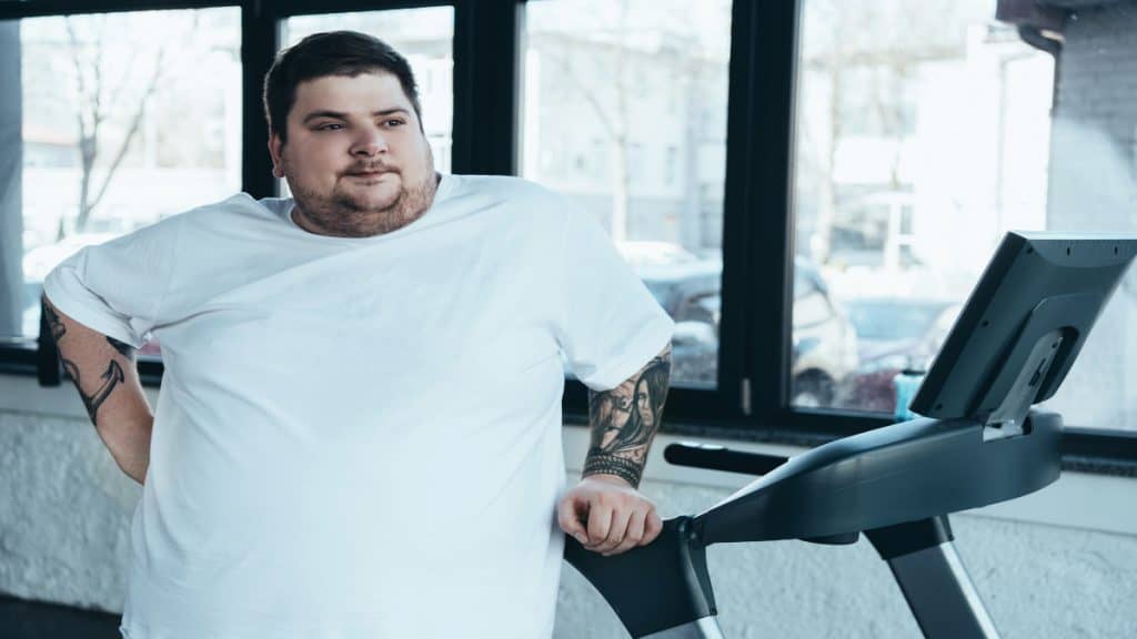 An overweight man with a 57 inch waist standing next to a treadmill
