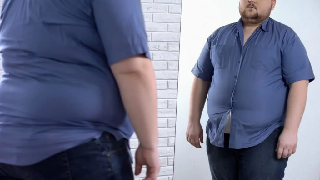 An obese man looking at his big 69 in waist in the mirror